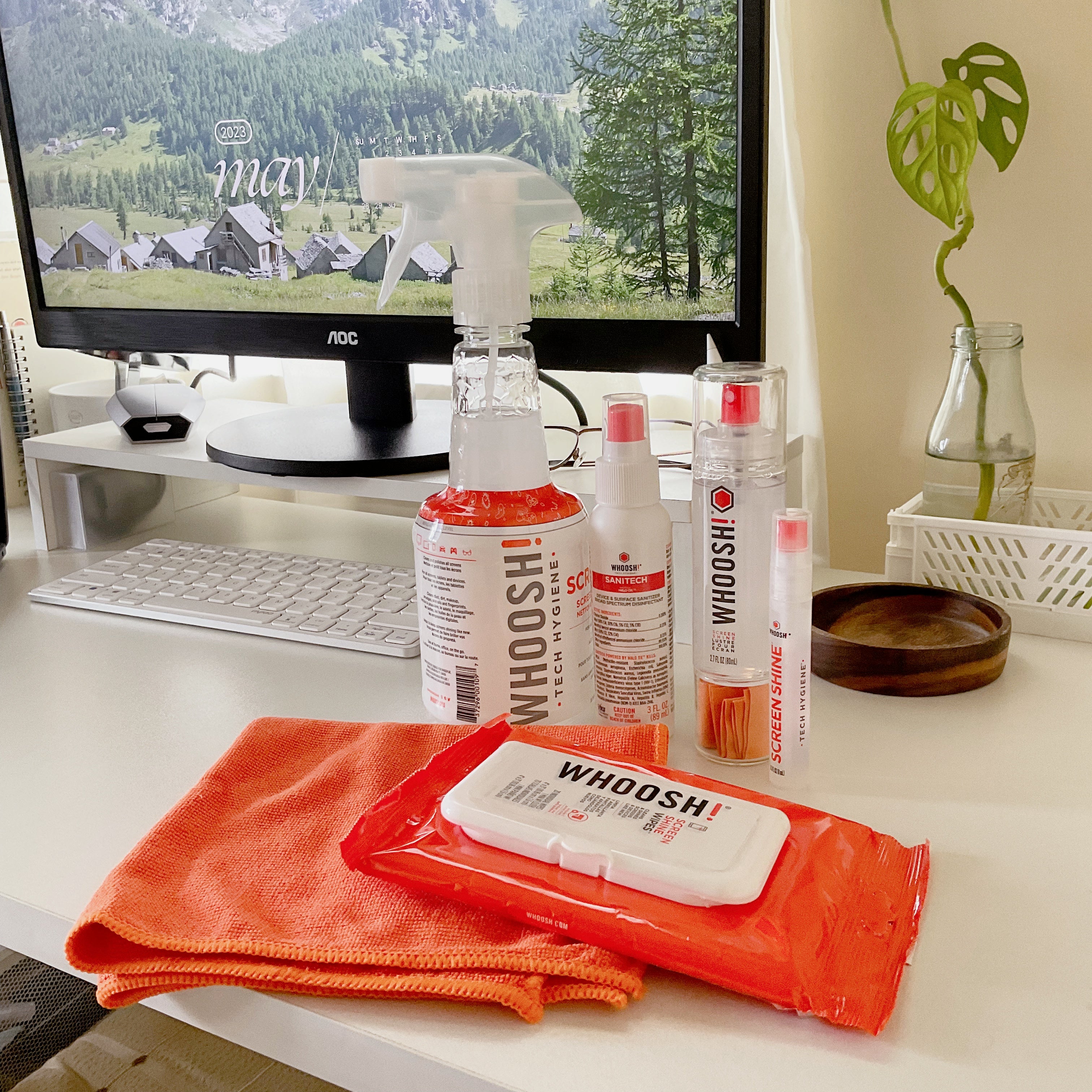 WHOOSH! screen cleaner spray, wipes and microfibre cloth on a bright desk beside a monitor and keyboard, showing a complete tech hygiene setup for clean, clear screens.
