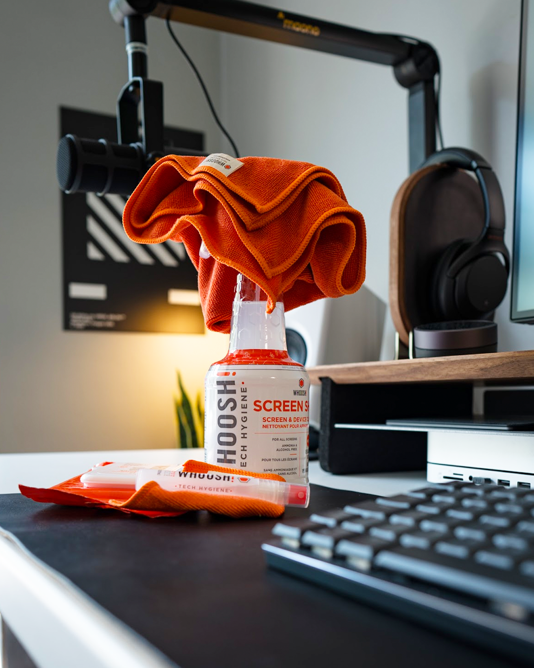 WHOOSH! screen cleaner and microfibre cloth on a modern desk beside a keyboard and headphones, showing easy everyday tech hygiene for screens and devices.