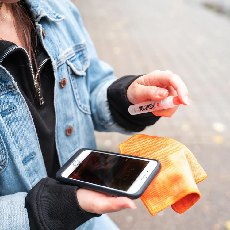 Person cleaning a smartphone screen with a WHOOSH! screen cleaner spray and orange microfibre cloth outdoors