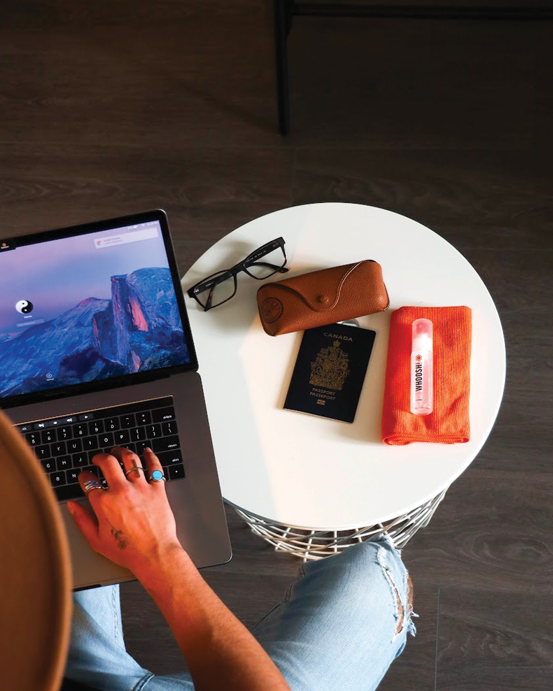 Person working on a laptop beside a small table with glasses, passport and WHOOSH! pocket screen cleaner and microfibre cloth, showing portable tech hygiene for travel and everyday life.